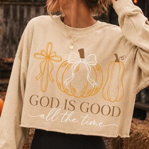 Puede incluir: Una sudadera corta de color beige con mangas largas que presenta un estampado gr&aacute;fico de una cruz, una calabaza con un lazo blanco y una calabaza alargada. El texto "GOD IS GOOD" est&aacute; impreso en marr&oacute;n, con "all the time" debajo en cursiva.