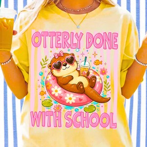 K&ouml;nnte beinhalten: Gelbes T-Shirt mit einer Grafik eines Otters mit herzf&ouml;rmiger Sonnenbrille und Schleife, der in einem rosa Schwimmring treibt. Der Text "OTTERLY DONE WITH SCHOOL" befindet sich &uuml;ber und unter der Otter-Illustration.