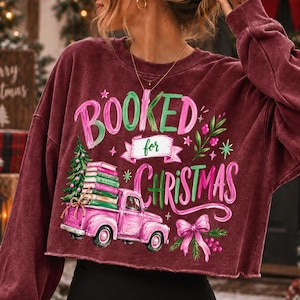 May include: A maroon long-sleeve sweatshirt featuring a graphic print with the text BOOKED for CHRISTMAS. The design includes a pink vintage truck carrying a stack of books and a Christmas tree, accented with green foliage and a pink bow.