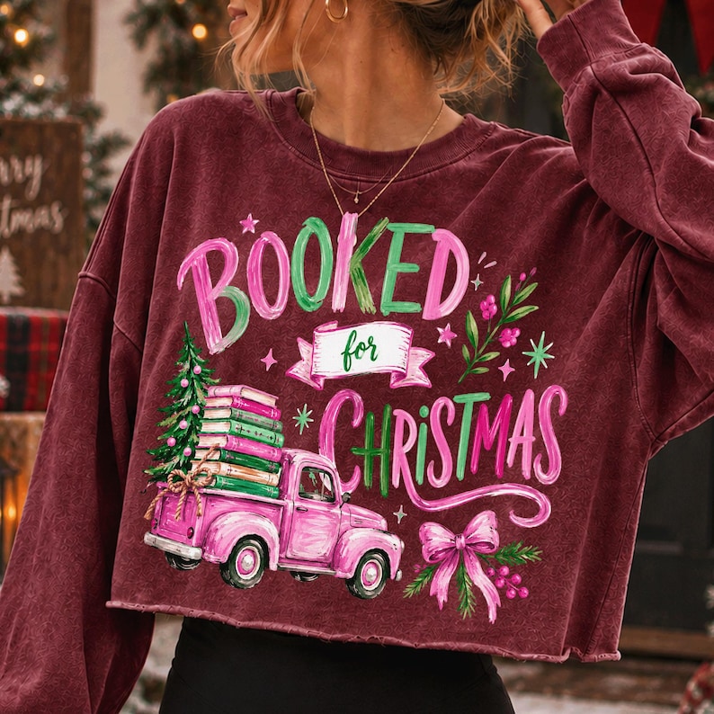 May include: A maroon long-sleeve sweatshirt featuring a graphic print with the text BOOKED for CHRISTMAS. The design includes a pink vintage truck carrying a stack of books and a Christmas tree, accented with green foliage and a pink bow.