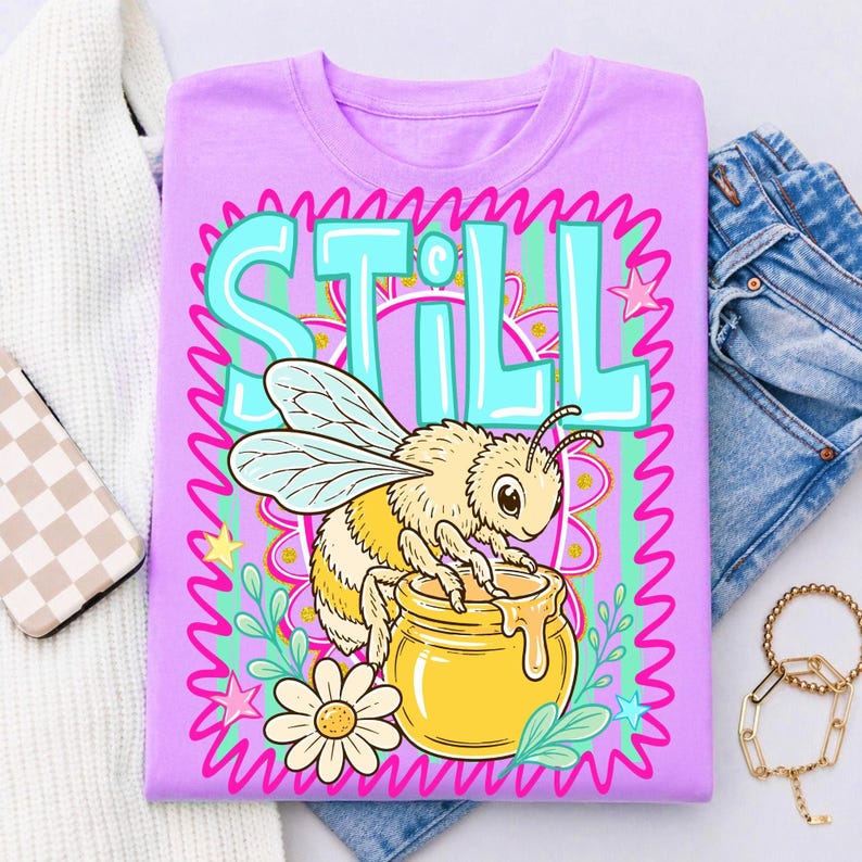 Op de afbeelding: Een lila T-shirt met een cartoon bijenontwerp. De bij, met gele en zwarte strepen, zit op een honingpot. Het woord "STILL" staat in turquoise, omringd door een bloemenpatroon.