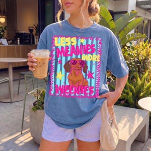 May include: A blue t-shirt featuring the phrase "LESS MEANIES MORE WEENIES" and a cartoon dachshund wearing sunglasses. The shirt has a faded appearance and is worn with striped shorts. The style is casual and playful.