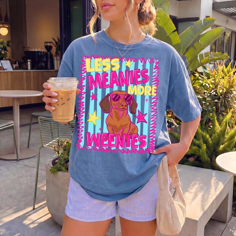 May include: A blue t-shirt featuring the phrase "LESS MEANIES MORE WEENIES" and a cartoon dachshund wearing sunglasses. The shirt has a faded appearance and is worn with striped shorts. The style is casual and playful.