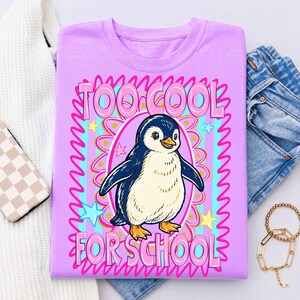 K&ouml;nnte beinhalten: Helllila T-Shirt mit einem Cartoon-Pinguin und dem Text "TOO COOL FOR SCHOOL" in Pink. Der Pinguin befindet sich in einem rosa Oval mit blauem und goldenem Rand, umgeben von Sternen.