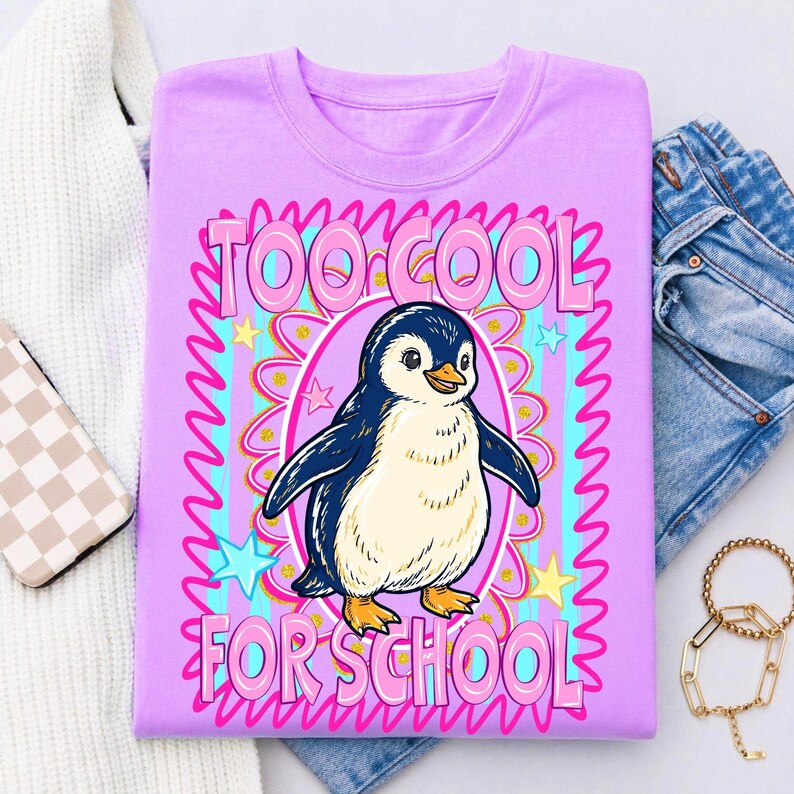 K&ouml;nnte beinhalten: Helllila T-Shirt mit einem Cartoon-Pinguin und dem Text "TOO COOL FOR SCHOOL" in Pink. Der Pinguin befindet sich in einem rosa Oval mit blauem und goldenem Rand, umgeben von Sternen.