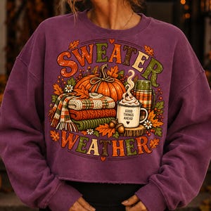 Puede incluir: Una sudadera de manga larga color ciruela con un estampado gr&aacute;fico que incluye el texto "SWEATER WEATHER". El dise&ntilde;o muestra una calabaza naranja grande, una pila de mantas a cuadros dobladas, un termo y una taza con "GOOD THINGS AHEAD".
