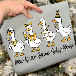 May include: A light gray fabric with a graphic of four cartoon geese celebrating. One goose wears sunglasses and holds a sparkler, another holds a champagne glass, and the last wears a party hat. Text reads "New Year Same Silly Goose."