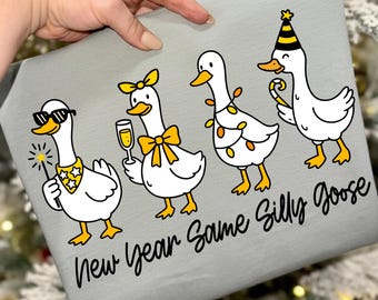 May include: A light gray fabric with a graphic of four cartoon geese celebrating. One goose wears sunglasses and holds a sparkler, another holds a champagne glass, and the last wears a party hat. Text reads "New Year Same Silly Goose."