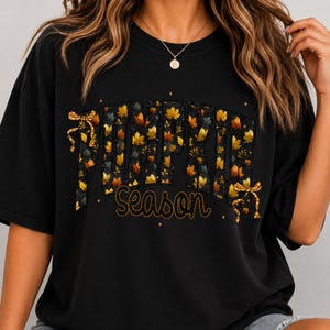 May include: A black short-sleeved t-shirt featuring the text PUMPKIN in large block letters filled with a pattern of autumn leaves. Below, the word Season is written in a cursive font. Two gold-colored bows are attached to the sides of the text.