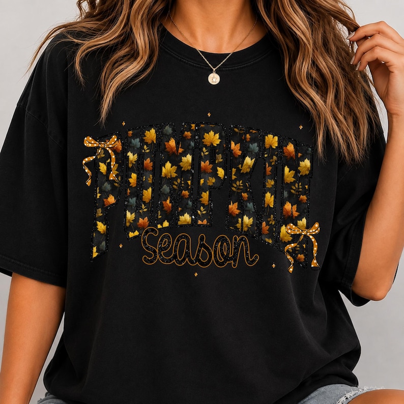 May include: A black short-sleeved t-shirt featuring the text PUMPKIN in large block letters filled with a pattern of autumn leaves. Below, the word Season is written in a cursive font. Two gold-colored bows are attached to the sides of the text.