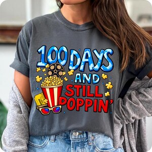 Puede incluir: Camiseta gris oscuro con el texto "100 Days and Still Poppin'". El dise&ntilde;o incluye una cubeta de palomitas de ma&iacute;z, una bobina de pel&iacute;cula, gafas 3D y una entrada de cine. El texto es azul y rojo.