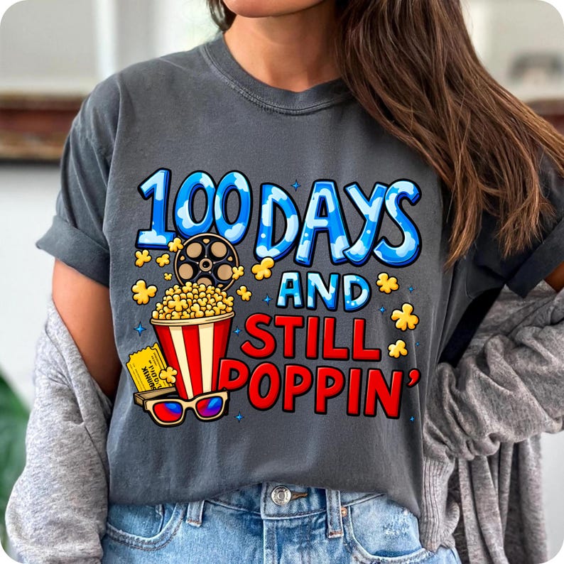 Puede incluir: Camiseta gris oscuro con el texto "100 Days and Still Poppin'". El dise&ntilde;o incluye una cubeta de palomitas de ma&iacute;z, una bobina de pel&iacute;cula, gafas 3D y una entrada de cine. El texto es azul y rojo.