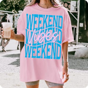 K&ouml;nnte beinhalten: Hellrosa T-Shirt mit dem Schriftzug "WEEKEND Vibes WEEKEND" in blau und rosa in einem wellenf&ouml;rmigen Design. Das T-Shirt ist oversized und hat kurze &Auml;rmel. Die Person tr&auml;gt Shorts und h&auml;lt ein Getr&auml;nk.