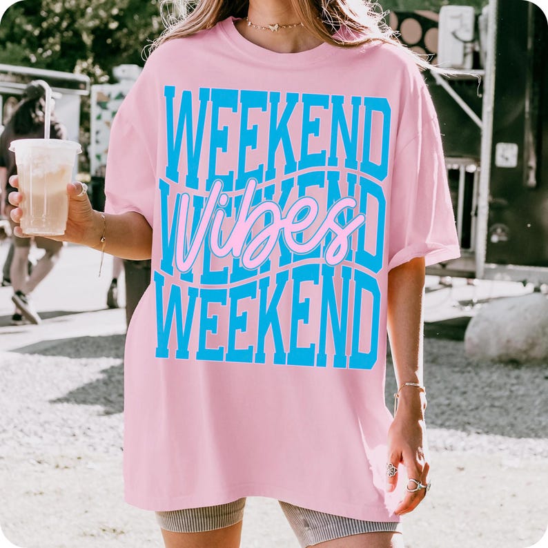 K&ouml;nnte beinhalten: Hellrosa T-Shirt mit dem Schriftzug "WEEKEND Vibes WEEKEND" in blau und rosa in einem wellenf&ouml;rmigen Design. Das T-Shirt ist oversized und hat kurze &Auml;rmel. Die Person tr&auml;gt Shorts und h&auml;lt ein Getr&auml;nk.