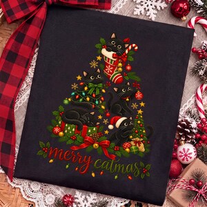 K&ouml;nnte beinhalten: Ein schwarzes T-Shirt mit einer Grafik von vier schwarzen Katzen in Form eines Weihnachtsbaums, verziert mit bunten Lichtern, Ornamenten und einem Weihnachtsstrumpf. Der Text "merry catmas" ist unten in Rot und Gr&uuml;n aufgedruckt.