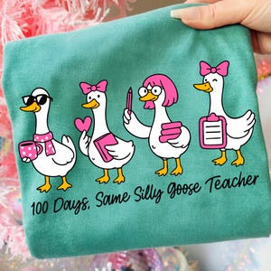 K&ouml;nnte beinhalten: T&uuml;rkisfarbenes T-Shirt mit vier Cartoon-G&auml;nsen, die rosa Schleifen tragen und verschiedene Gegenst&auml;nde halten. Die G&auml;nse sind mit Brillen und anderen Accessoires ausgestattet. Der Text lautet "100 Days. Same Silly Goose Teacher."