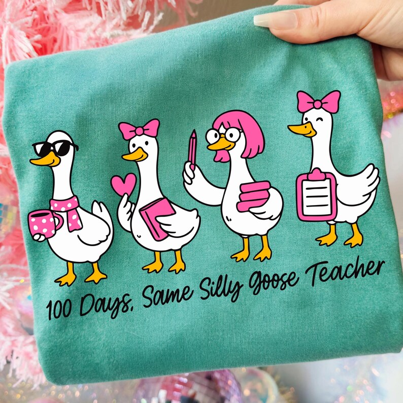 K&ouml;nnte beinhalten: T&uuml;rkisfarbenes T-Shirt mit vier Cartoon-G&auml;nsen, die rosa Schleifen tragen und verschiedene Gegenst&auml;nde halten. Die G&auml;nse sind mit Brillen und anderen Accessoires ausgestattet. Der Text lautet "100 Days. Same Silly Goose Teacher."