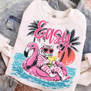 K&ouml;nnte beinhalten: Wei&szlig;es Sweatshirt mit einem Aufdruck eines Tigers mit herzf&ouml;rmiger Sonnenbrille, der auf einem rosa Flamingo-Schwimmer entspannt. Das Wort "Easy" steht in rosa Schrift &uuml;ber dem Tiger. Palmen und ein Sonnenuntergang sind im Hintergrund.