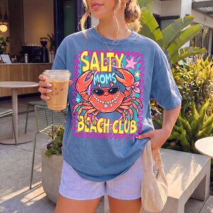 May include: A blue t-shirt featuring the text "Salty Moms Beach Club" and a cartoon crab wearing sunglasses and a pearl necklace. The crab is orange with pink and yellow accents. The shirt is worn with striped shorts.