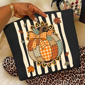 May include: A black sweatshirt featuring a central graphic of a patchwork pumpkin with an orange gingham bow. The pumpkin is surrounded by vertical cream stripes and scattered autumn leaves. The design includes various patterns like polka dots.