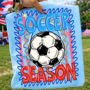 Puede incluir: Camiseta azul claro con las palabras "SOCCER SEASON" en azul y rojo, y un gr&aacute;fico de bal&oacute;n de f&uacute;tbol en blanco y negro. El dise&ntilde;o est&aacute; rodeado por un borde ondulado azul con estrellas y detalles dorados.