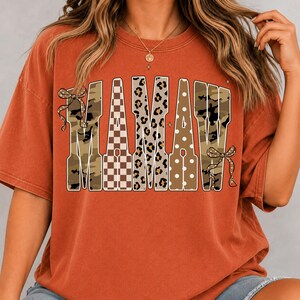 May include: A rust-coloured short-sleeve t-shirt featuring the word MAMAW in large block letters. Each letter has a different pattern including camouflage, brown and white checkerboard, leopard print, and white polka dots, with bows on the ends.