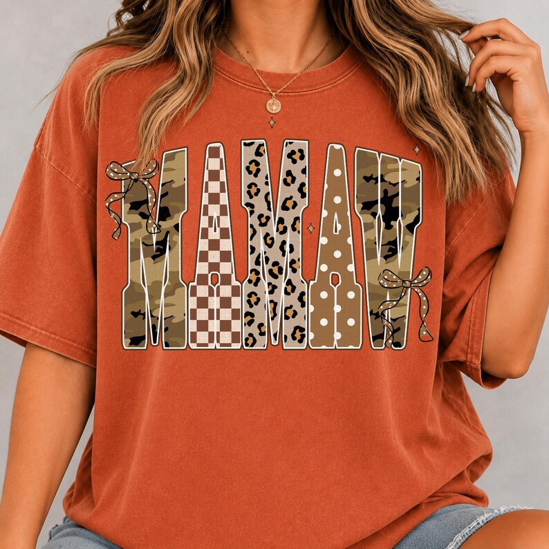 May include: A rust-coloured short-sleeve t-shirt featuring the word MAMAW in large block letters. Each letter has a different pattern including camouflage, brown and white checkerboard, leopard print, and white polka dots, with bows on the ends.