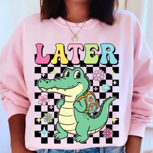 K&ouml;nnte beinhalten: Hellrosa Sweatshirt mit dem Wort "LATER" in bunten Buchstaben &uuml;ber einem Cartoon-Alligator mit Rucksack. Der Alligator ist gr&uuml;n und gelb, vor einem schwarz-wei&szlig; karierten Hintergrund mit Blumen- und Sternakzenten.