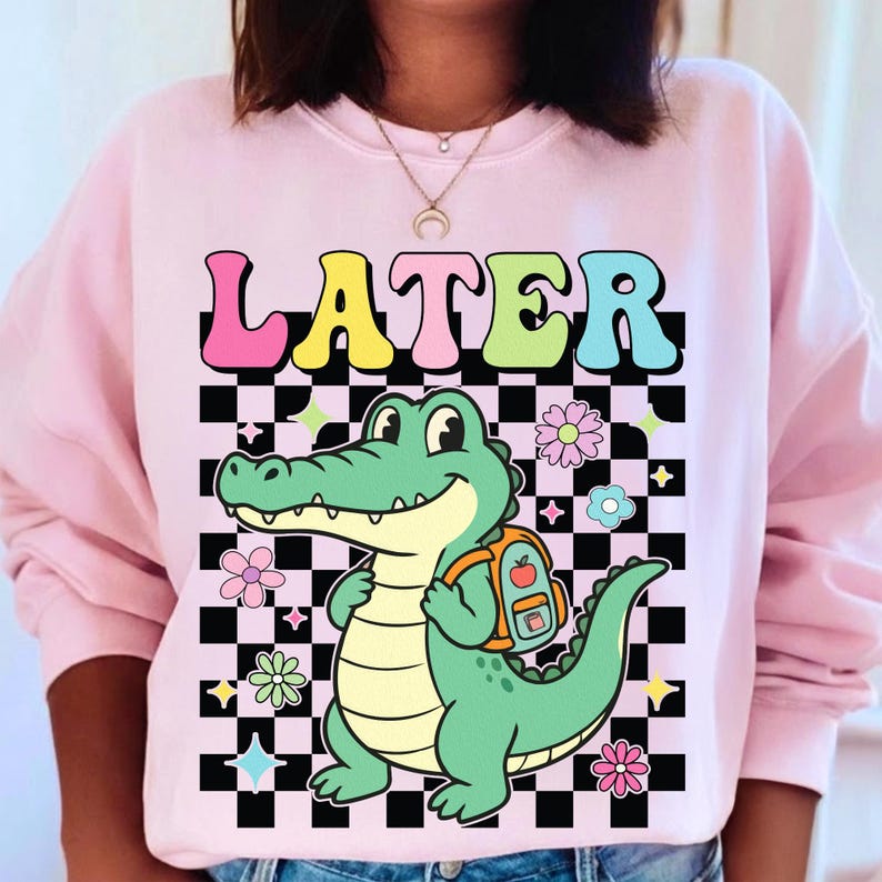 K&ouml;nnte beinhalten: Hellrosa Sweatshirt mit dem Wort "LATER" in bunten Buchstaben &uuml;ber einem Cartoon-Alligator mit Rucksack. Der Alligator ist gr&uuml;n und gelb, vor einem schwarz-wei&szlig; karierten Hintergrund mit Blumen- und Sternakzenten.