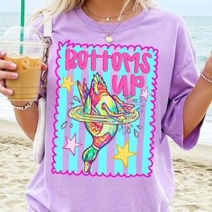May include: A lilac t-shirt with the phrase "BOTTOMS UP" in a pink and blue graphic. The design shows a duck diving into water, with stars and a wavy border. The shirt has short sleeves and a relaxed fit.