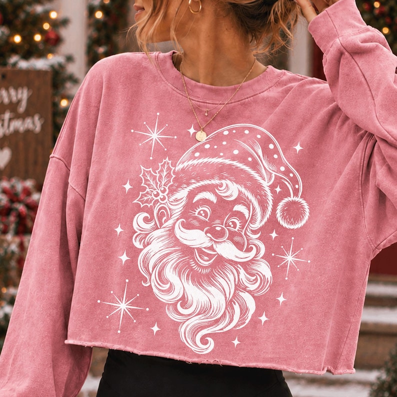 May include: A dusty rose long-sleeve cropped sweatshirt featuring a white graphic print of a smiling Santa Claus face wearing a hat with a pom-pom, accented by holly leaves and starburst shapes. The garment has a crew neckline and raw hem.