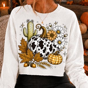 Puede incluir: Una sudadera blanca de manga larga con un estampado gr&aacute;fico de una calabaza con manchas de vaca, una calabaza de cuadros amarillos, un cactus verde, hojas de oto&ntilde;o marrones, bellotas, tallos de trigo y margaritas blancas y amarillas.