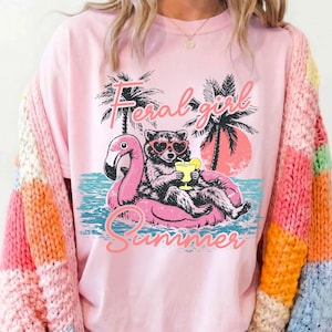 Pode incluir: Camiseta rosa claro com um desenho de um guaxinim usando &oacute;culos de sol em forma de cora&ccedil;&atilde;o, flutuando em um flutuador de flamingo rosa. O texto "Feral Girl Summer" est&aacute; acima e abaixo da imagem. Palmeiras e um p&ocirc;r do sol no fundo.