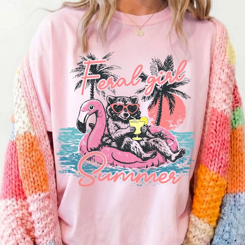 Pode incluir: Camiseta rosa claro com um desenho de um guaxinim usando &oacute;culos de sol em forma de cora&ccedil;&atilde;o, flutuando em um flutuador de flamingo rosa. O texto "Feral Girl Summer" est&aacute; acima e abaixo da imagem. Palmeiras e um p&ocirc;r do sol no fundo.