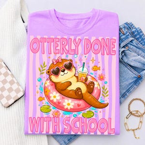 K&ouml;nnte beinhalten: Helllila T-Shirt mit dem Text "OTTERLY DONE WITH SCHOOL" in Pink. Die Grafik zeigt einen Cartoon-Otter mit herzf&ouml;rmiger Sonnenbrille, der auf einem rosa Schwimmring schwimmt und ein Getr&auml;nk h&auml;lt. Der Hintergrund hat rosa und lila Streifen.