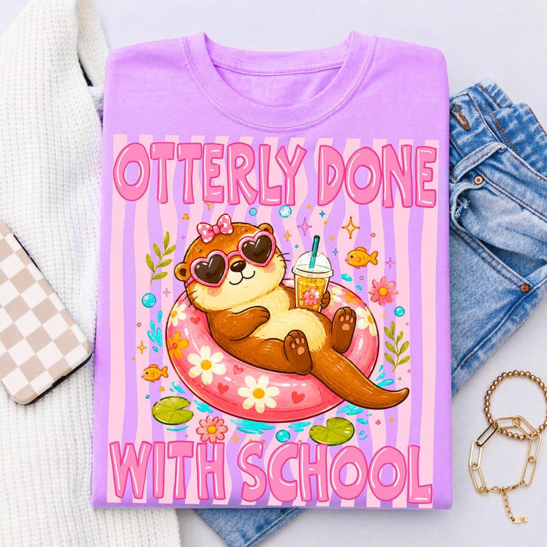 K&ouml;nnte beinhalten: Helllila T-Shirt mit dem Text "OTTERLY DONE WITH SCHOOL" in Pink. Die Grafik zeigt einen Cartoon-Otter mit herzf&ouml;rmiger Sonnenbrille, der auf einem rosa Schwimmring schwimmt und ein Getr&auml;nk h&auml;lt. Der Hintergrund hat rosa und lila Streifen.