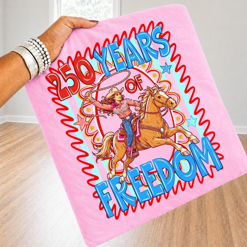 May include: Pink blanket with a graphic of a cowgirl riding a horse, roping a lasso. The blanket has the text "250 YEARS OF FREEDOM" in red and blue. The blanket is held by a person.