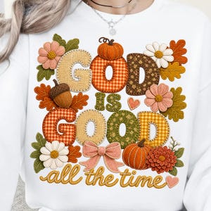 Pu&ograve; includere: Una felpa bianca con un design grafico che riporta il testo "GOD IS GOOD all the time". Le lettere sono riempite con vari motivi autunnali tra cui quadri, fiori e texture, circondate da zucche, foglie e fiori.