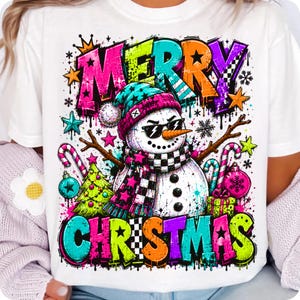 Puede incluir: Una camiseta blanca con un gr&aacute;fico colorido de un mu&ntilde;eco de nieve con gafas de sol, una bufanda de cuadros y un gorro. El mu&ntilde;eco est&aacute; rodeado de &aacute;rboles de Navidad, bastones de caramelo y adornos. El texto MERRY CHRISTMAS est&aacute; en negrita.