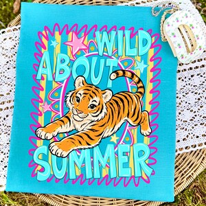 K&ouml;nnte beinhalten: Eine t&uuml;rkisfarbene Tragetasche mit einem farbenfrohen Design. Die Tasche zeigt einen Zeichentiger mit orangefarbenen und schwarzen Streifen und die Worte "WILD ABOUT SUMMER" in T&uuml;rkis, Rosa und Gelb. Das Design wird von einem rosa-gelben Rahmen umrandet.