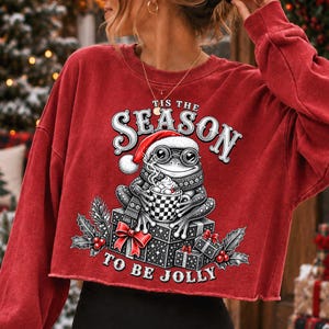 Puede incluir: Una sudadera roja corta de manga larga con el gr&aacute;fico de una rana con gorro de Pap&aacute; Noel y gafas, sosteniendo una taza de cacao sobre cajas de regalo. El texto dice Tis The Season To Be Jolly, rodeado de acebo y bayas.
