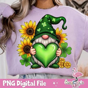 Op de afbeelding: Een paars sweatshirt met een groene gnome die een groen hart vasthoudt. De gnome draagt ​​een groene hoed met klavertjes en een witte baard. De gnome wordt omringd door zonnebloemen en klavertjes.