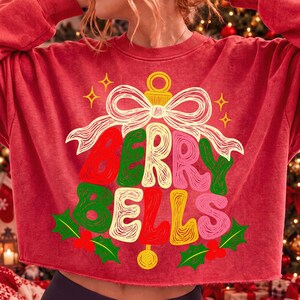 May include: A red crewneck sweatshirt featuring a graphic of a Christmas ornament shaped like a bell with a bow on top. The text BERRY BELLS is written in a stylized, multi-colored font. Green holly leaves and red berries are at the bottom.