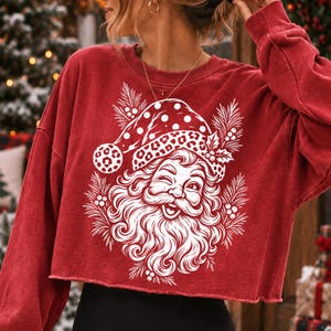 K&ouml;nnte beinhalten: Ein rotes, lang&auml;rmeliges, bauchfreies Sweatshirt mit einem wei&szlig;en Grafikdruck eines zwinkernden Weihnachtsmanns, der eine M&uuml;tze mit Leopardenmuster tr&auml;gt, umgeben von Tannennadeln und Stechpalmenbeeren, mit einem unges&auml;umten Saum.