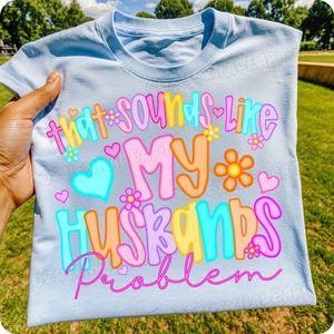 K&ouml;nnte beinhalten: Hellblaues T-Shirt mit dem Schriftzug "That sounds like MY HUSBANDS Problem" in bunten Buchstaben. Das Design umfasst Herzen und Blumen. Das Shirt ist aus einem weichen Material.