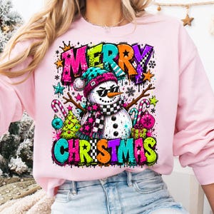 Puede incluir: Una sudadera rosa claro con un gr&aacute;fico vibrante de un mu&ntilde;eco de nieve con gafas de sol, un gorro azul verdoso y una bufanda a cuadros. El dise&ntilde;o incluye el texto MERRY CHRISTMAS en letras estilo grafiti, rodeado de estrellas, bastones de caramelo y regalos.
