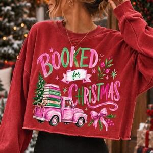 May include: A red long-sleeve sweatshirt featuring a graphic print with the text "BOOKED for CHRISTMAS". The design includes a pink pickup truck carrying a stack of books and a Christmas tree, accented with a pink bow and green foliage.