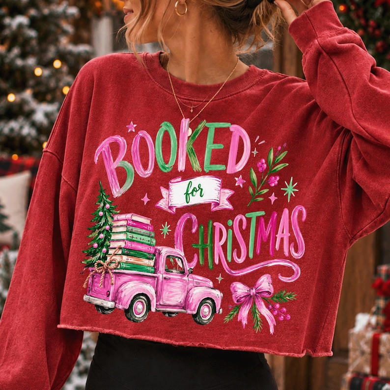 May include: A red long-sleeve sweatshirt featuring a graphic print with the text "BOOKED for CHRISTMAS". The design includes a pink pickup truck carrying a stack of books and a Christmas tree, accented with a pink bow and green foliage.
