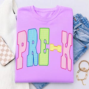May include: A light purple t-shirt featuring the text PRE-K in large, blocky letters. Each letter has a vertical striped pattern in alternating colors of pink, blue, green, and yellow. The shirt has a crew neckline and short sleeves.