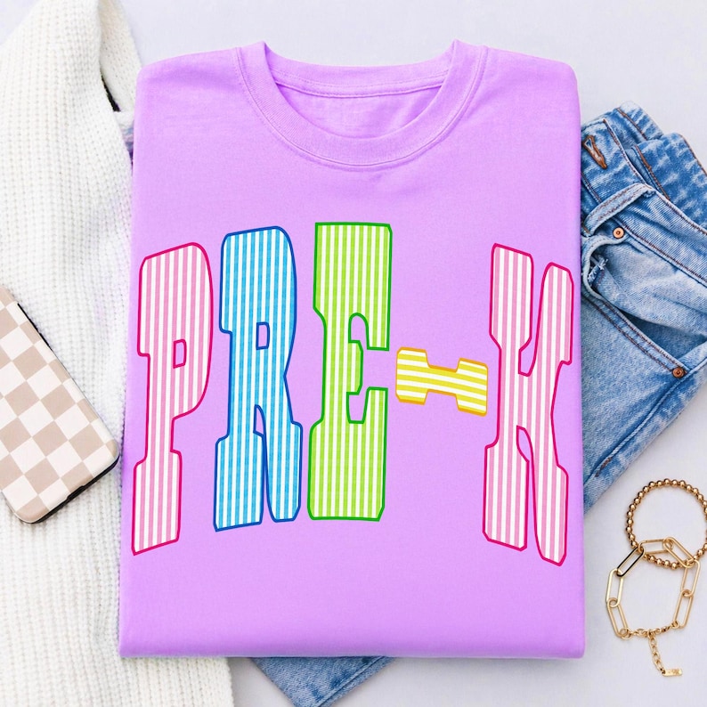 May include: A light purple t-shirt featuring the text PRE-K in large, blocky letters. Each letter has a vertical striped pattern in alternating colors of pink, blue, green, and yellow. The shirt has a crew neckline and short sleeves.